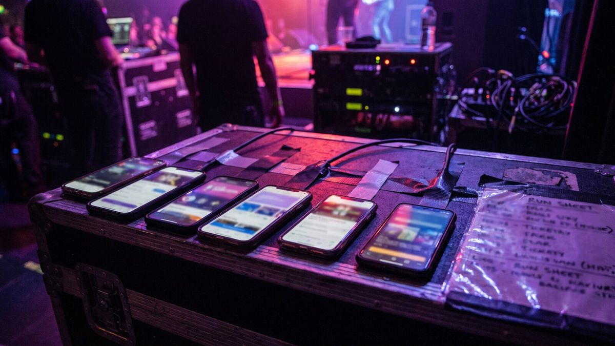 Six different phones lined up on a flight case backstage, each showing a different ticketing app's event page, gaffer tape and a laminated run sheet around them.