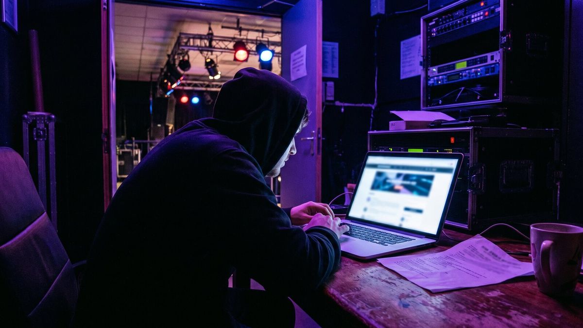 A promoter at a laptop in a small venue office, event page on screen, a printed run sheet and a coffee beside it, venue lights half-rigged in the background.